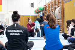 Eine Trainerin zeigt exemplarisch an einer Turnerin mehreren Personen eine Übung.
